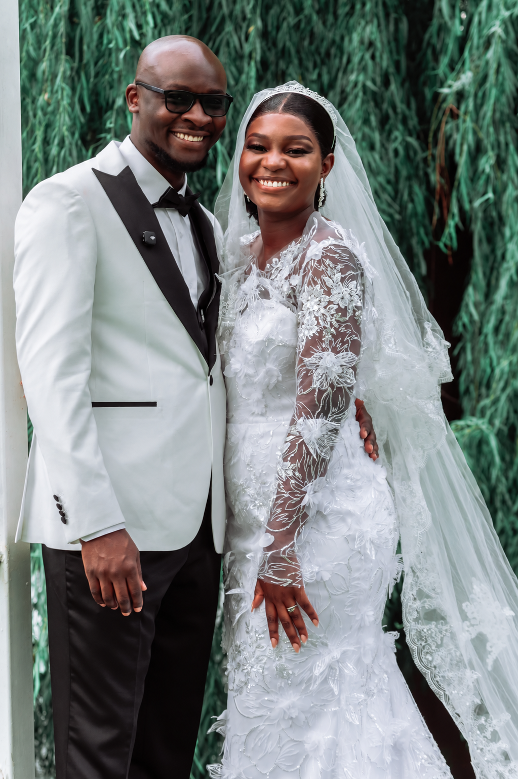 Smiling bride and groom in a white jacket by a tree